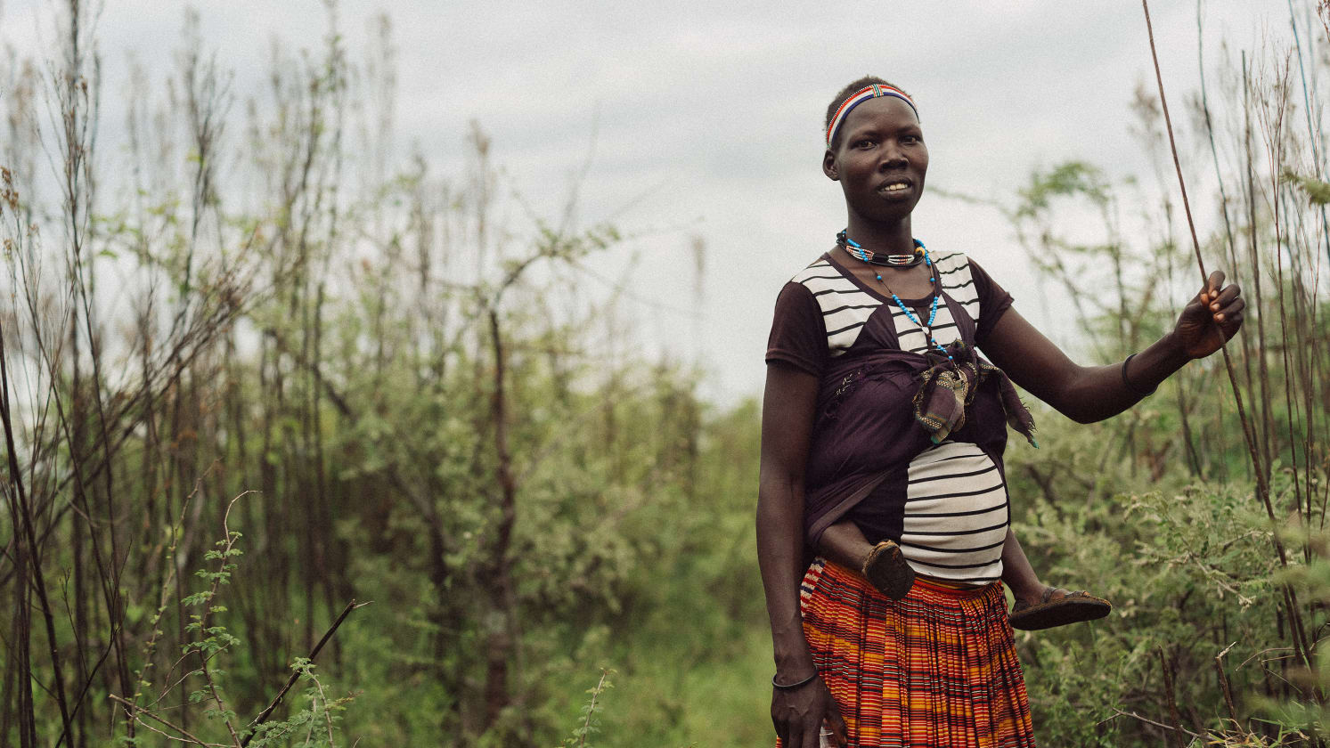 A woman with a beaded headband and necklace carries a baby in a front wrap, wearing a striped top and a plaid skirt, standing amidst green bushes and holding a stick.