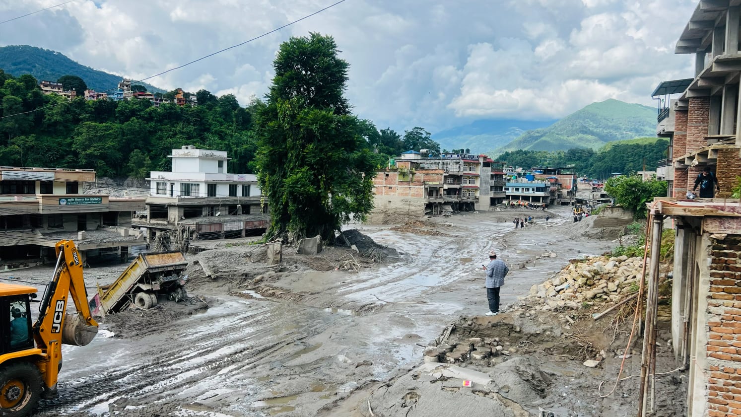 A wide view of a muddy landscape featuring damaged multi-storey buildings and construction equipment. A yellow JCB digger is on the left, with a partially submerged truck behind it. Several people are visible in the muddy expanse. In the distance are green hills and mountains under a cloudy sky.