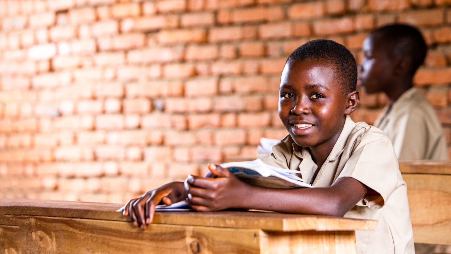 A smiling child sitting at a wooden desk with a brick background