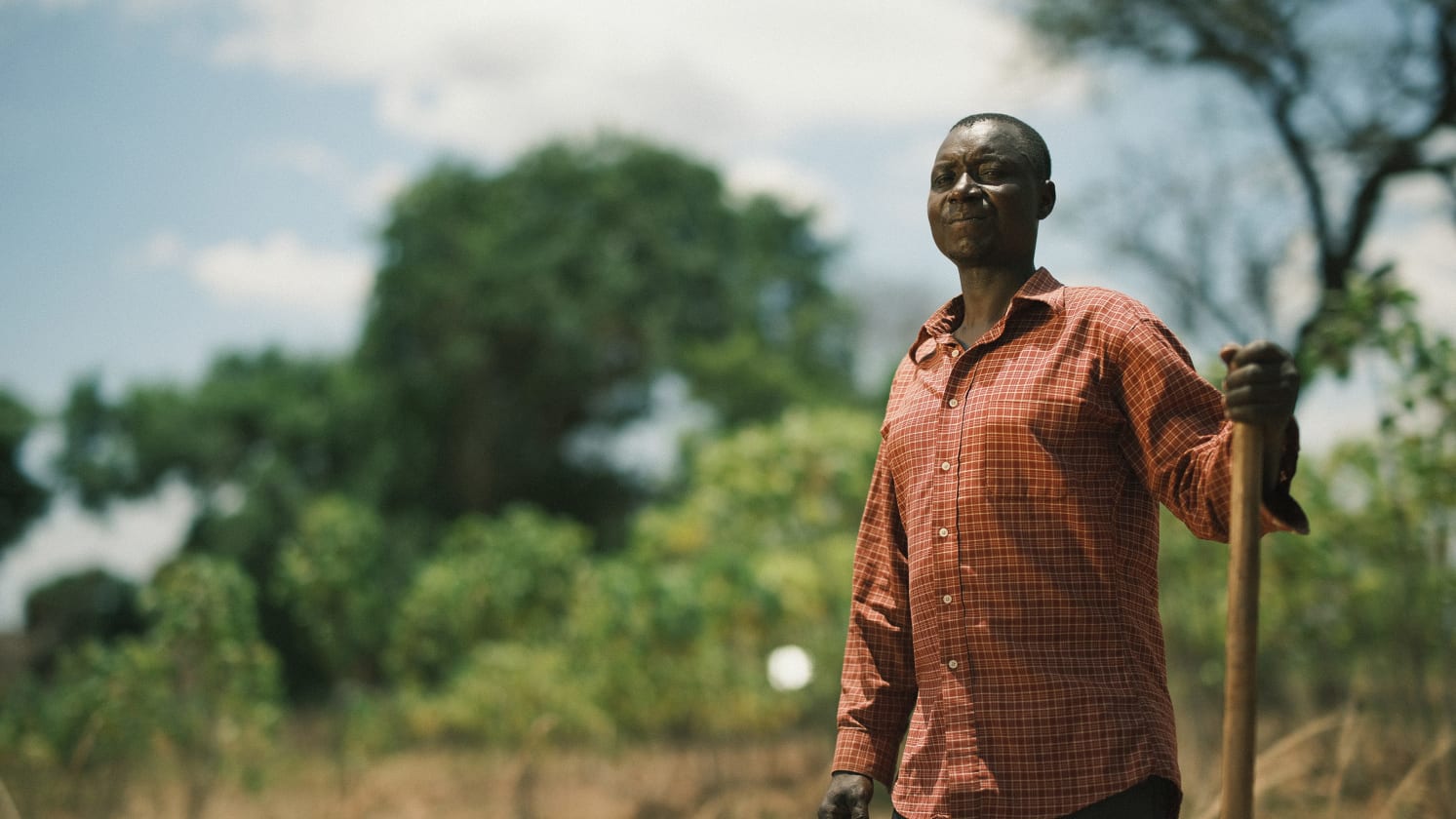 A man (named Isaac) standing in a field, leaning on a tool in his left hand. 
