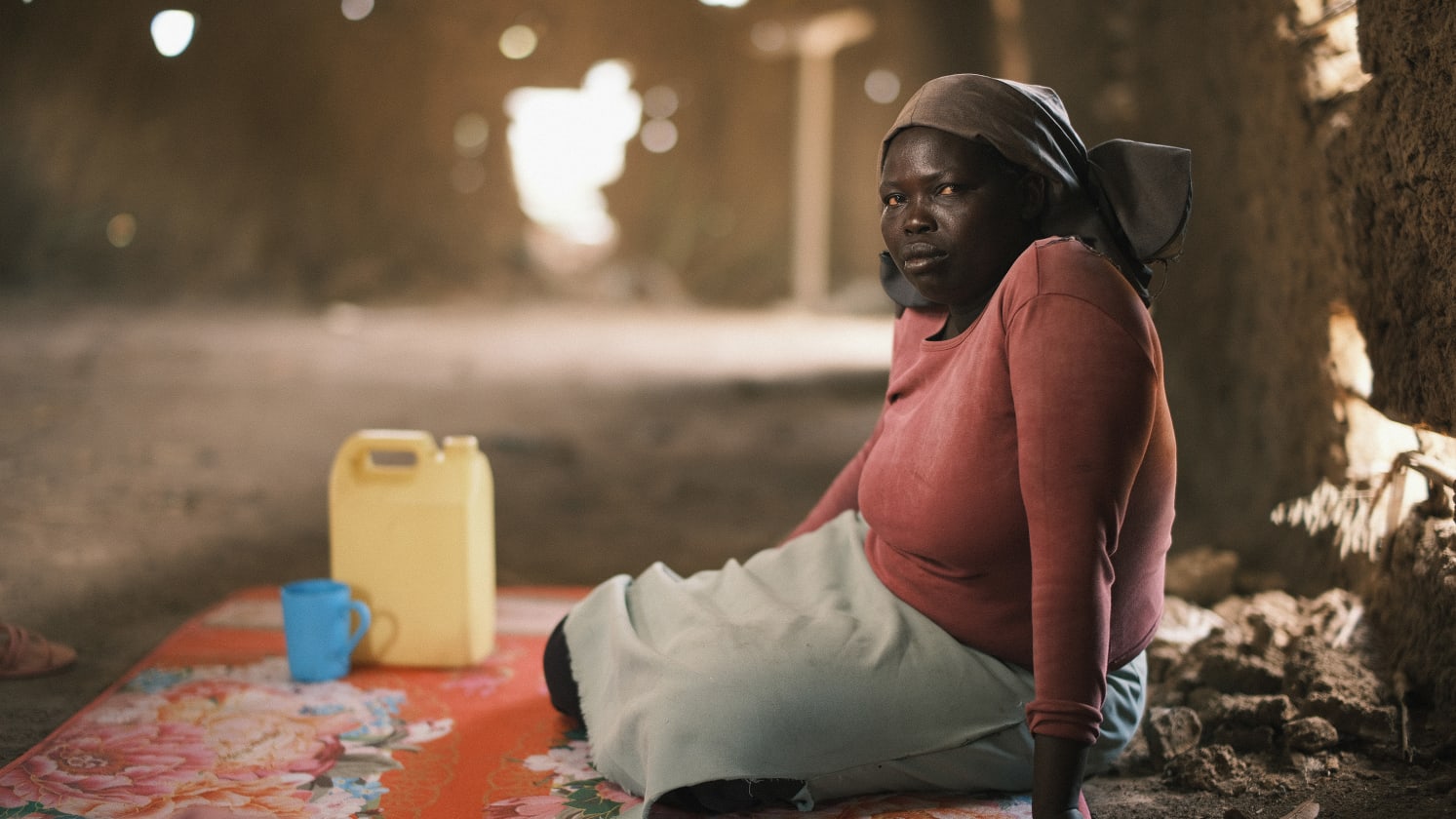 A woman sits inside a rustic building with a water jug and cup on a floral mat.
