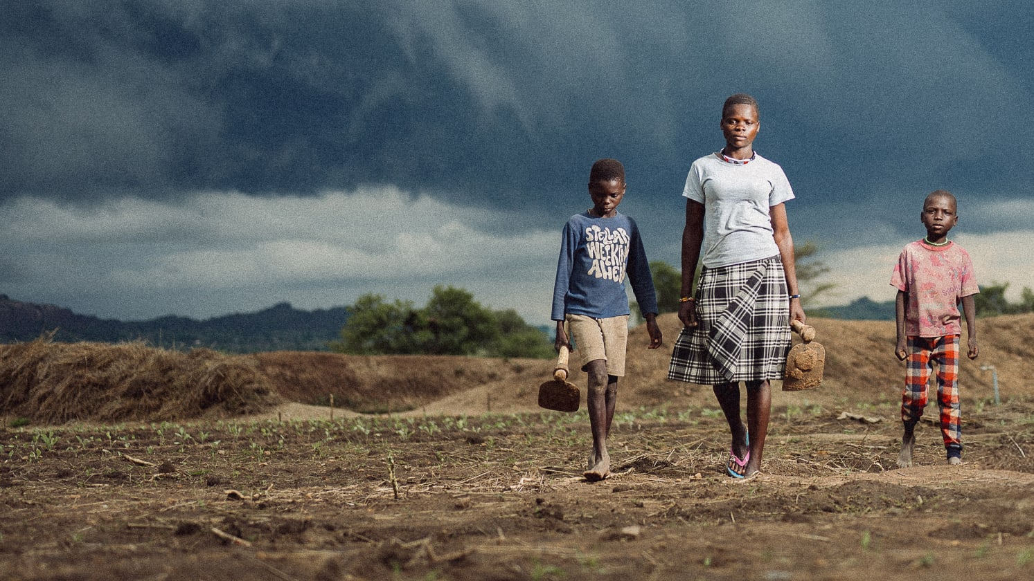 A young woman walks along a field with young children either side of her. Behind are dark rain clouds.