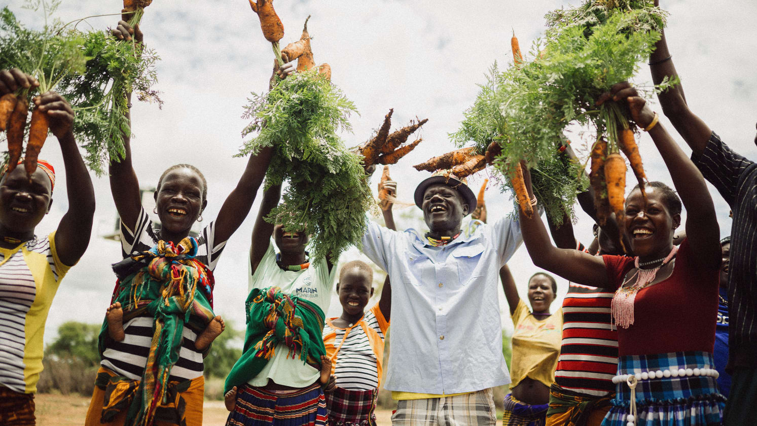 Joyful group holding up freshly harvested carrots with green tops under a bright sky.