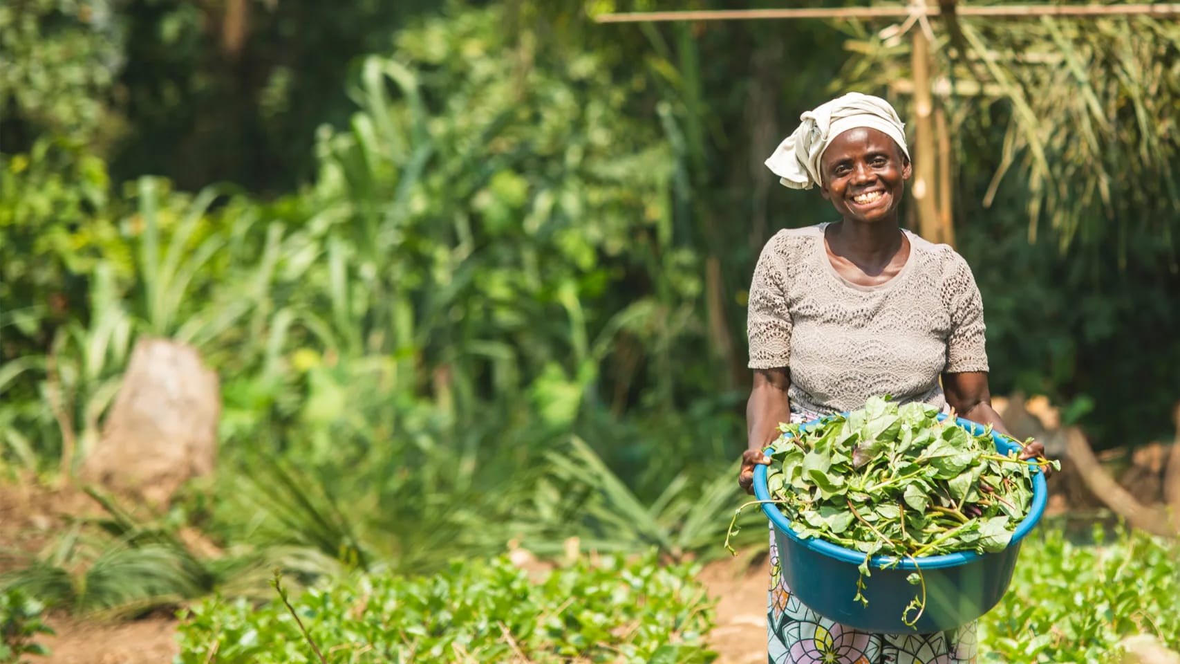 A lady outside, smiling, carrying a container of freshly-picked vegetables