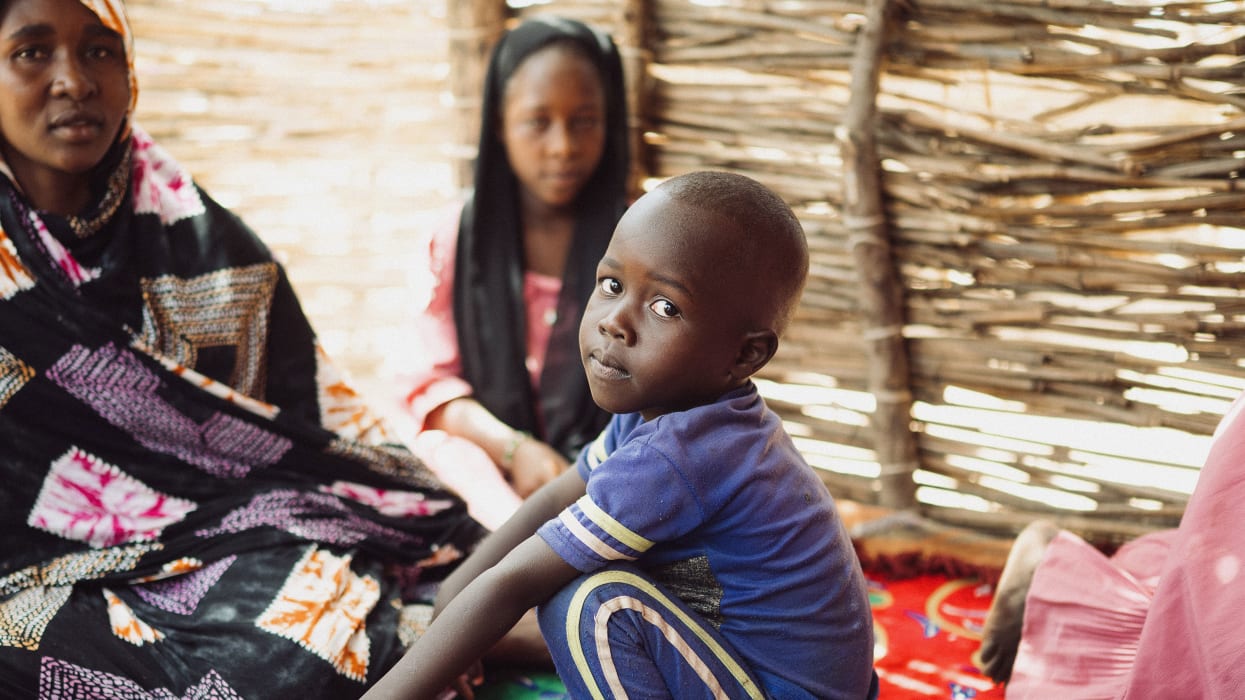 A young child in a blue tshirt looks at the camera, two women in traditional dress sit behind him.