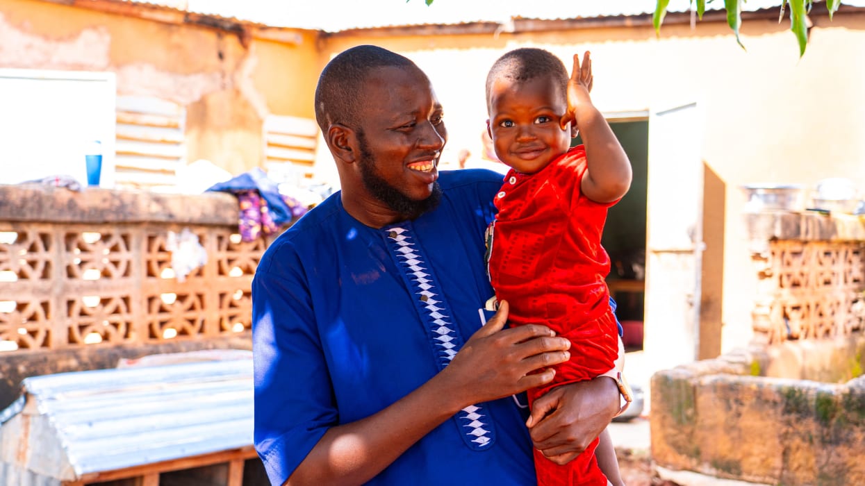 A man in a blue tunic, Gauthier, smiling warmly as he holds his young son. The child is waving at the camera. They are standing outdoors in a sunlit courtyard.