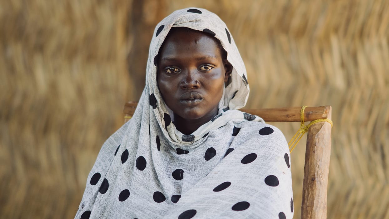 Young woman with dark skin and a nose piercing, wearing a white headscarf with black polka dots, looking directly forward. She sits in a simple wooden chair with a blurred, natural background.