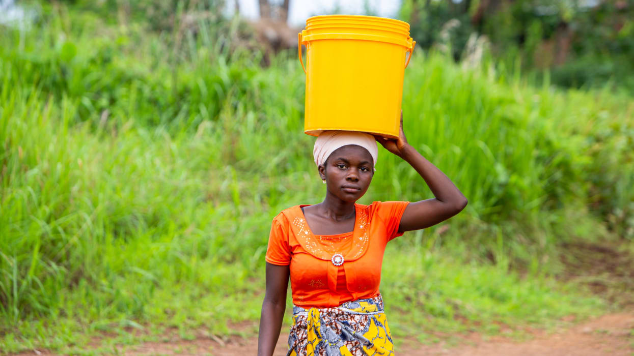 A woman looks ahead, holding a yellow bucket on top of her head with one arm.
