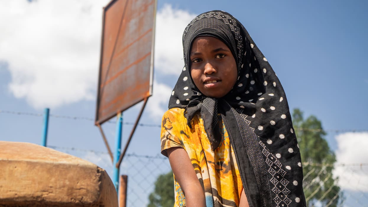 Portrait of a young woman in a hijab and yellow shirt, outdoors on a sunny day.