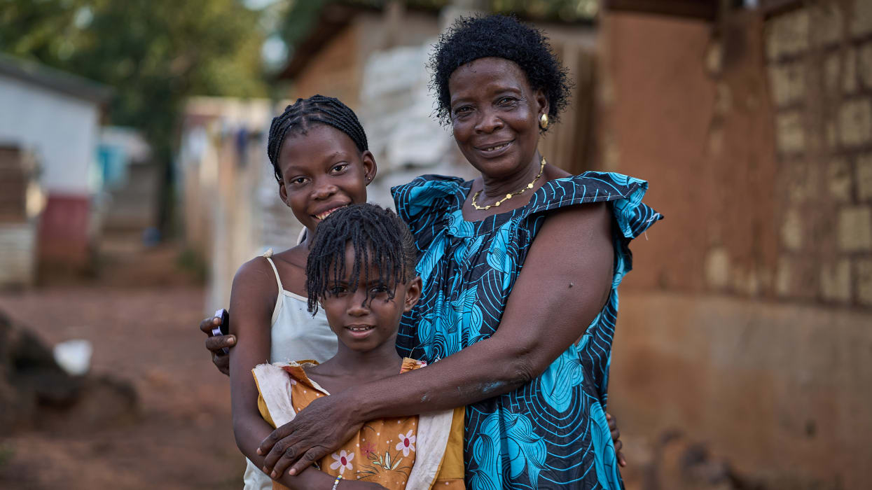 A woman in a blue dress stands smiling with her arms around two young girls.