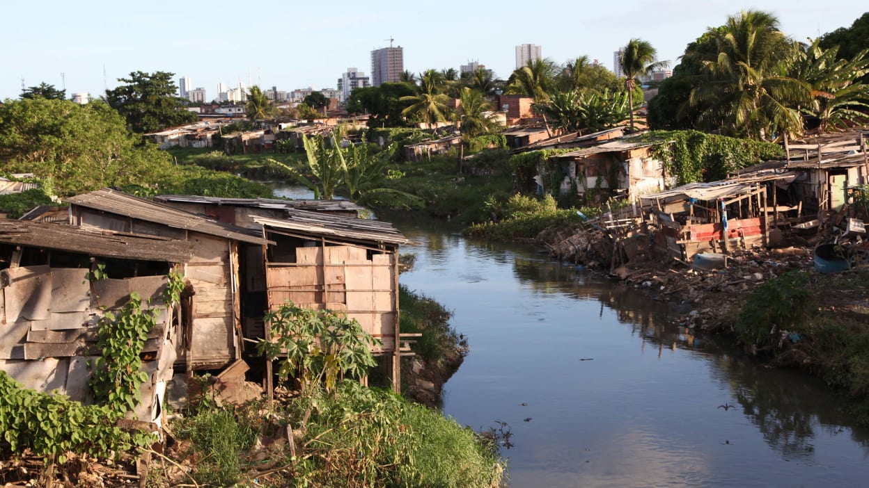 Habitações em uma favela, construídas nas duas margens de um pequeno rio no Brasil, com a cidade ao longe.