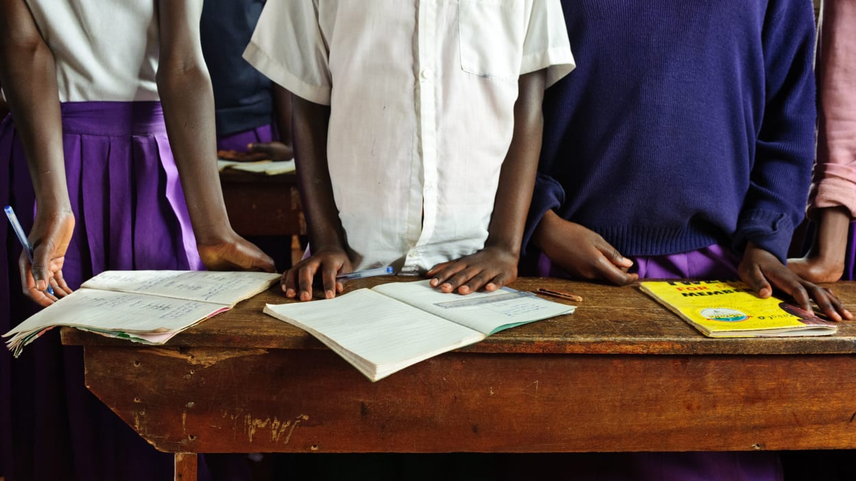 A close-up of the torsos of three African children standing side by side at a desk, with pens in hand and books open.