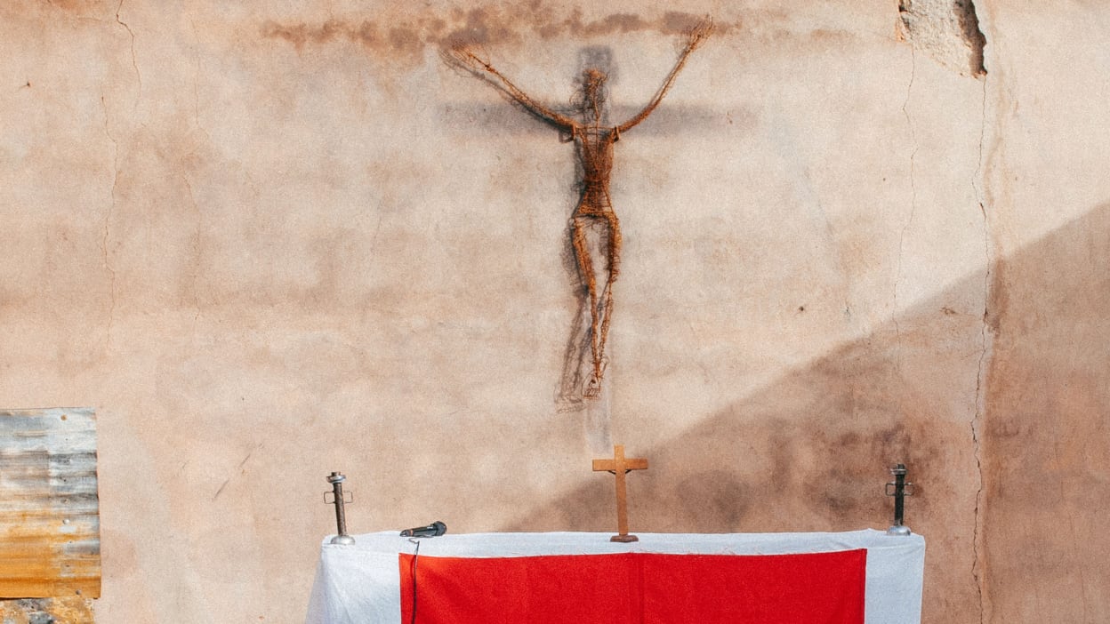 A representation of Christ on the cross constructed from rusty wire hangs on a bare, water-damaged wall in a Nigerian church.