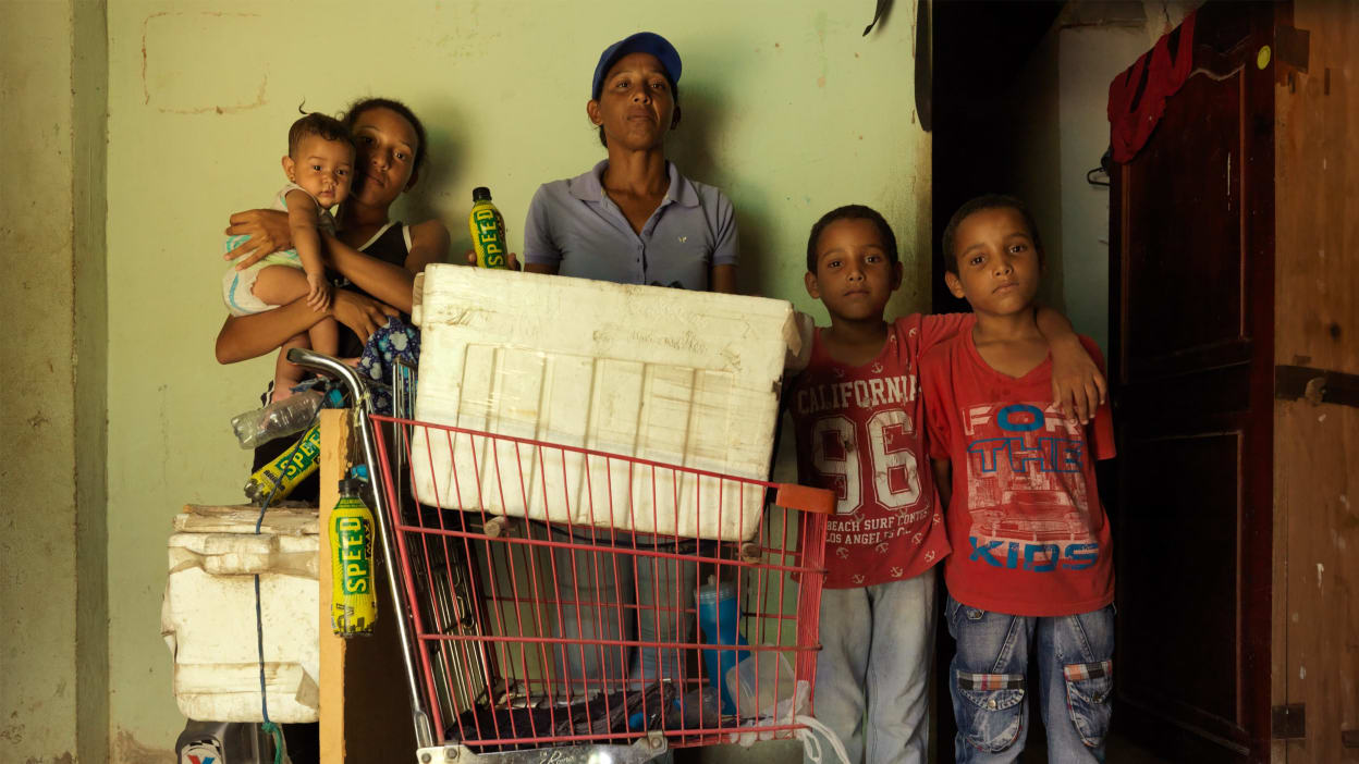 A woman and her four children all stand around a trolley with various items in it, the eldest child is holding a baby.