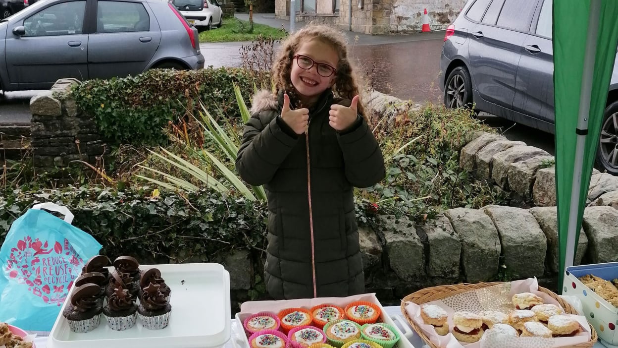 Amelie giving two thumbs up next to a stall with cakes, books and toys.