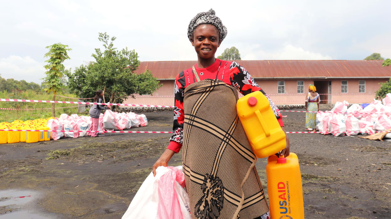 A woman carrying two jerry cans of clean water and a bag containing basic household items.