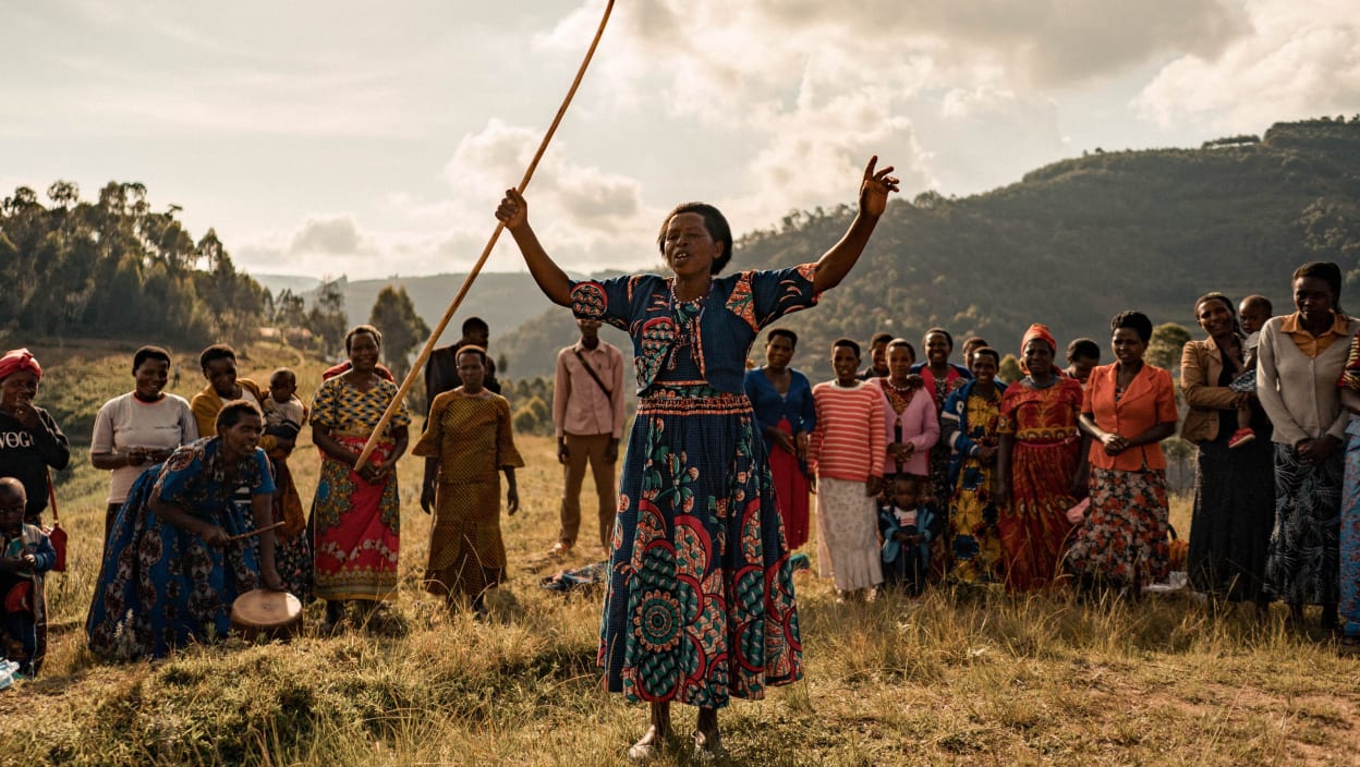 A woman dressed in a colourful patterened dress, lifts her arms up with a stick in one hand. Behind her stand a large group of people, one person is bent over and banging a small drum. 