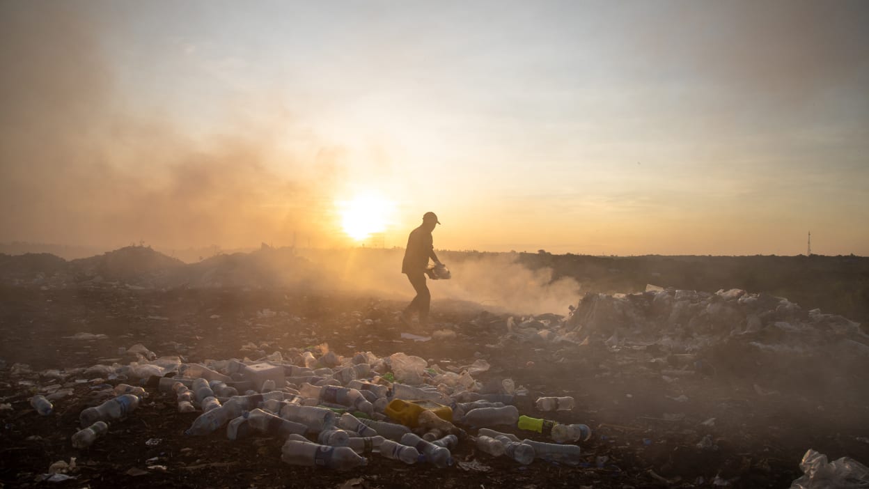 A dark silhouette of a person standing amongst plastic waste lying on the ground. Behind him is smoke billowing up into the air. 