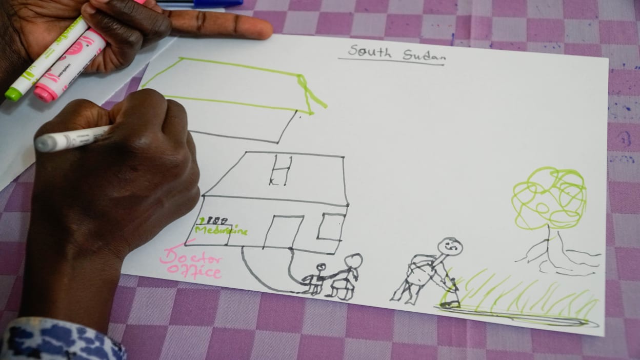 A peacebuilding workshop attendee from South Sudan draws a picture of what a peaceful community could look like. She has drawn a person farming happily in a field and people going to see a doctor in a health clinic.