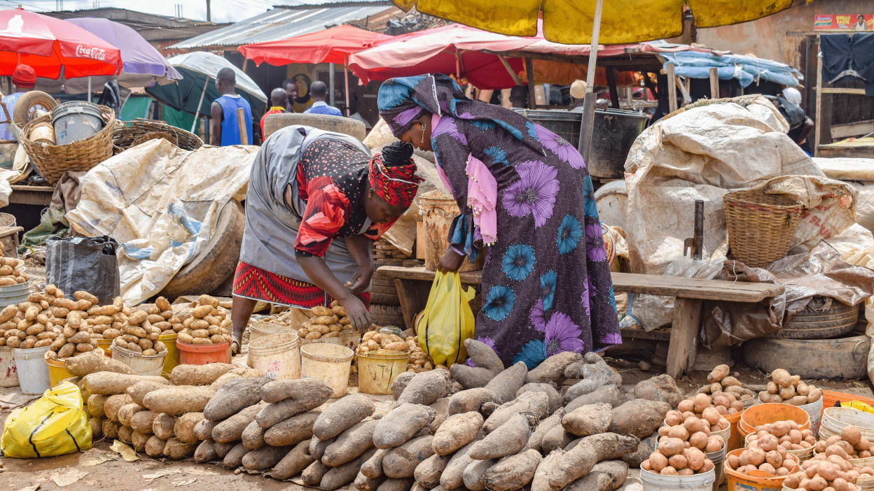 Two women in brightly-coloured dresses sell potatoes and other root vegetables in a street market in Nigeria where inflation of food prices reached 23 per cent in September 2022.