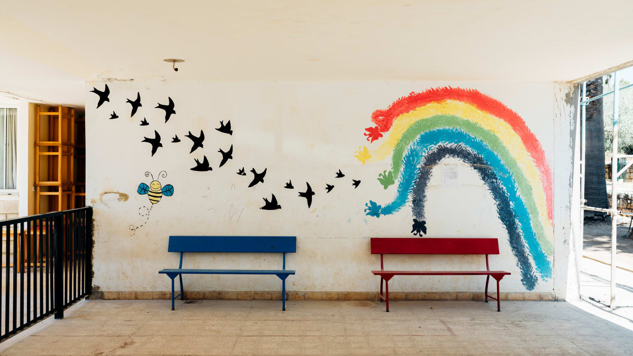 In a courtyard a wall has a painting of a rainbow and birds swooping next to it. In front of the painted wall are two benches.