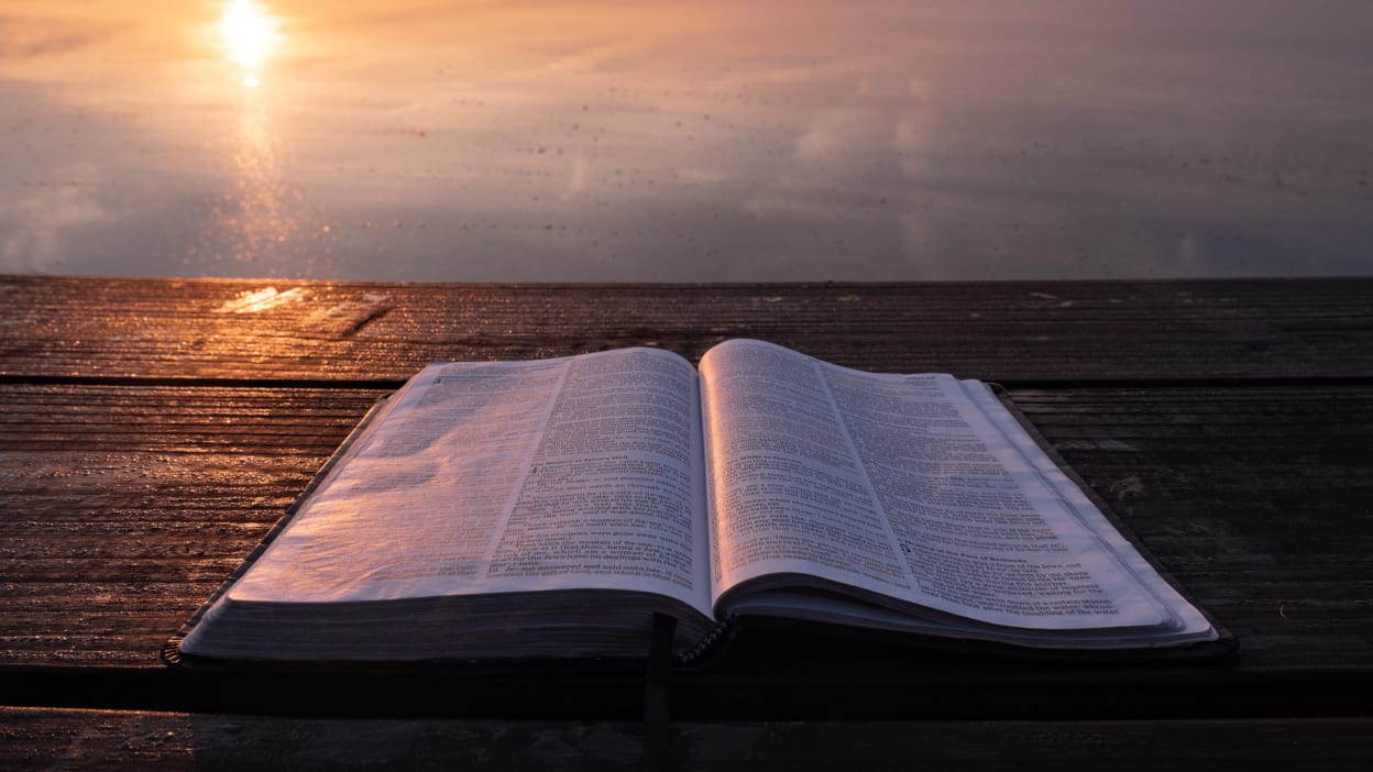 A Bible lies open on a table with a view of water stretching to the horizon as the sun sets.