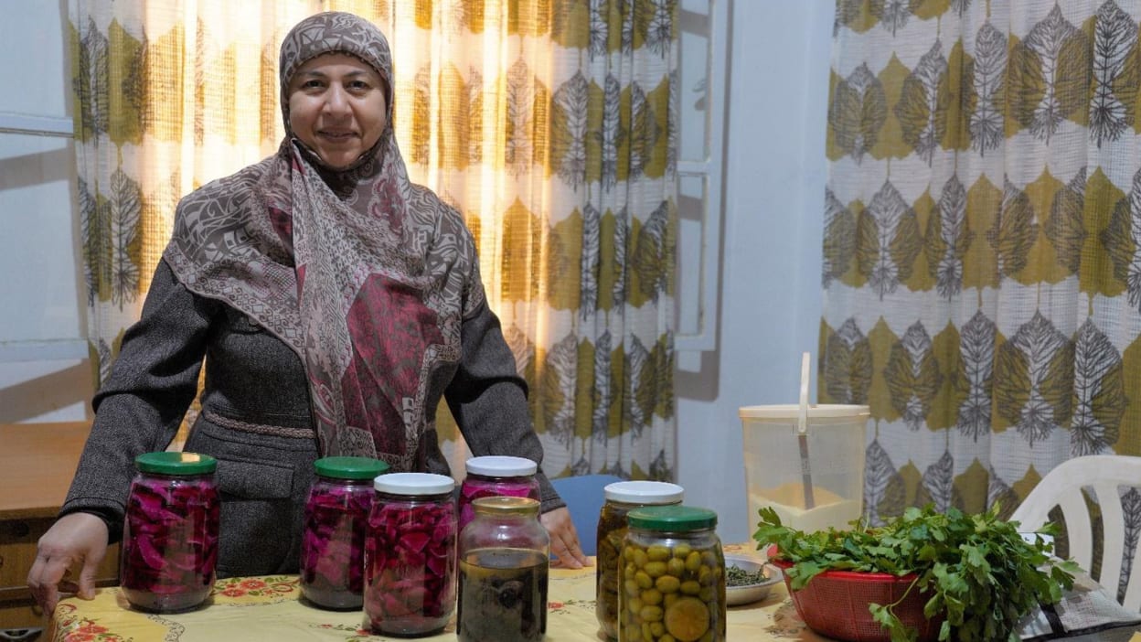 Lena, pictured wearing a headscarf and dress, shows some of the jars of products she prepares and sells to make a living.