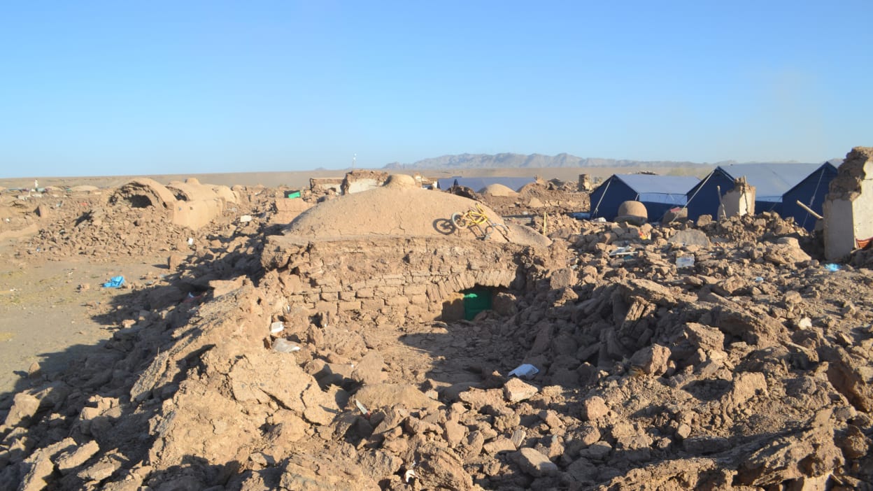 A child's bicycle sits on top of a pile of brown rubble that used to be a home. The dark blue tents shown in the background are providing some shelter for people who have been left homeless by the earthquakes. 