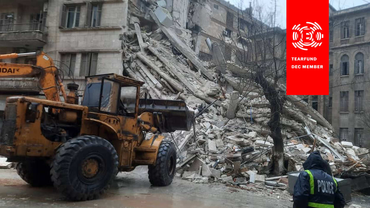 A large orange digger stands in front of a half-destroyed building, ready to start to clear away the rubble.