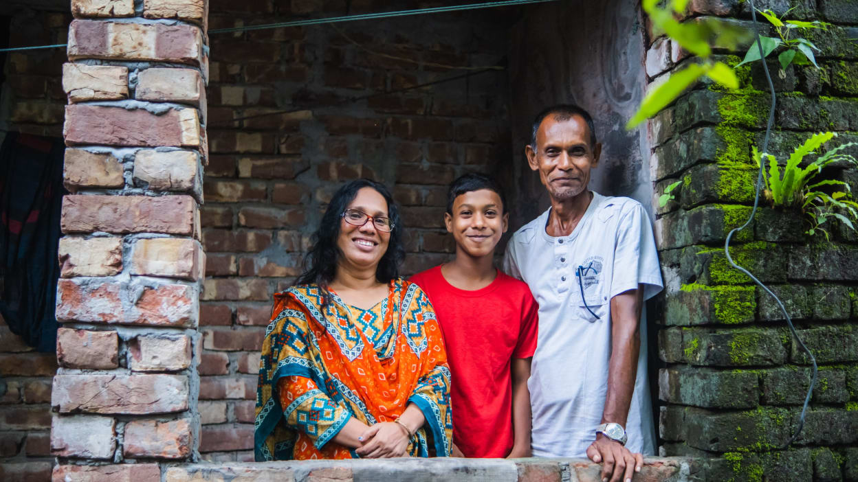 Shushanto, a man in a pale T-shirt, and Rheka, a woman with long, dark hair and wearing glasses and a brightly coloured outfit, stand smiling with one of their sons stood between them. 
