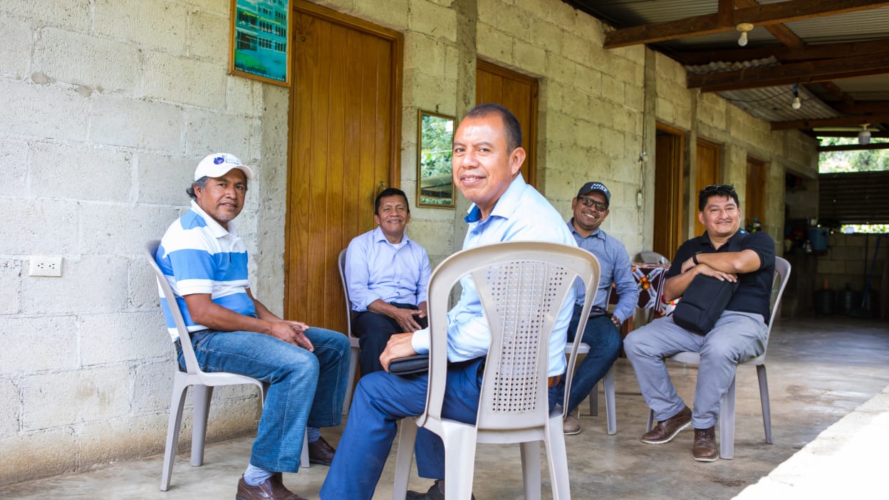A group of five men sit in a circle on plastic chairs under a shelter. 
