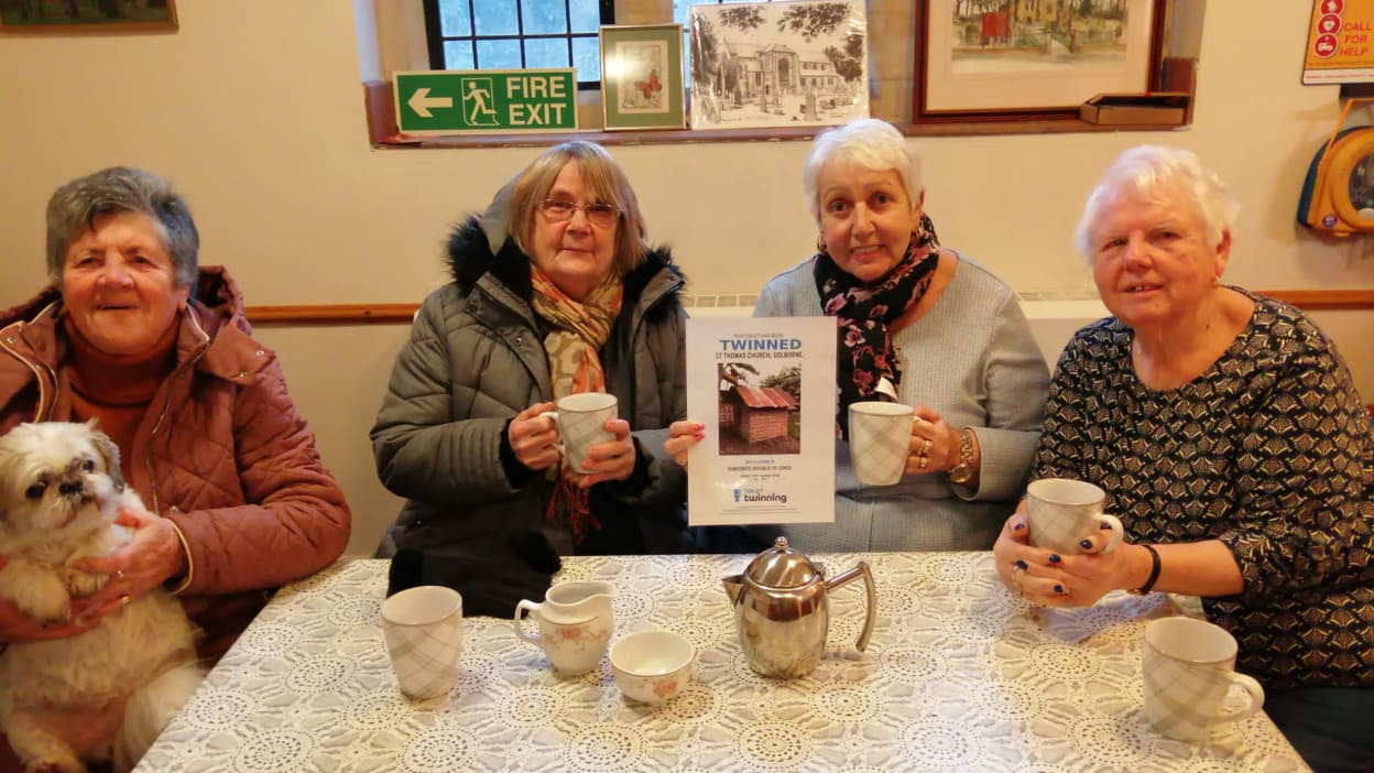 The four women, ages ranging from 70 to 82, sit around a table with tea cups and a saucer and their Toilet Twinning certificate. The woman on the far left holds a small white dog. 