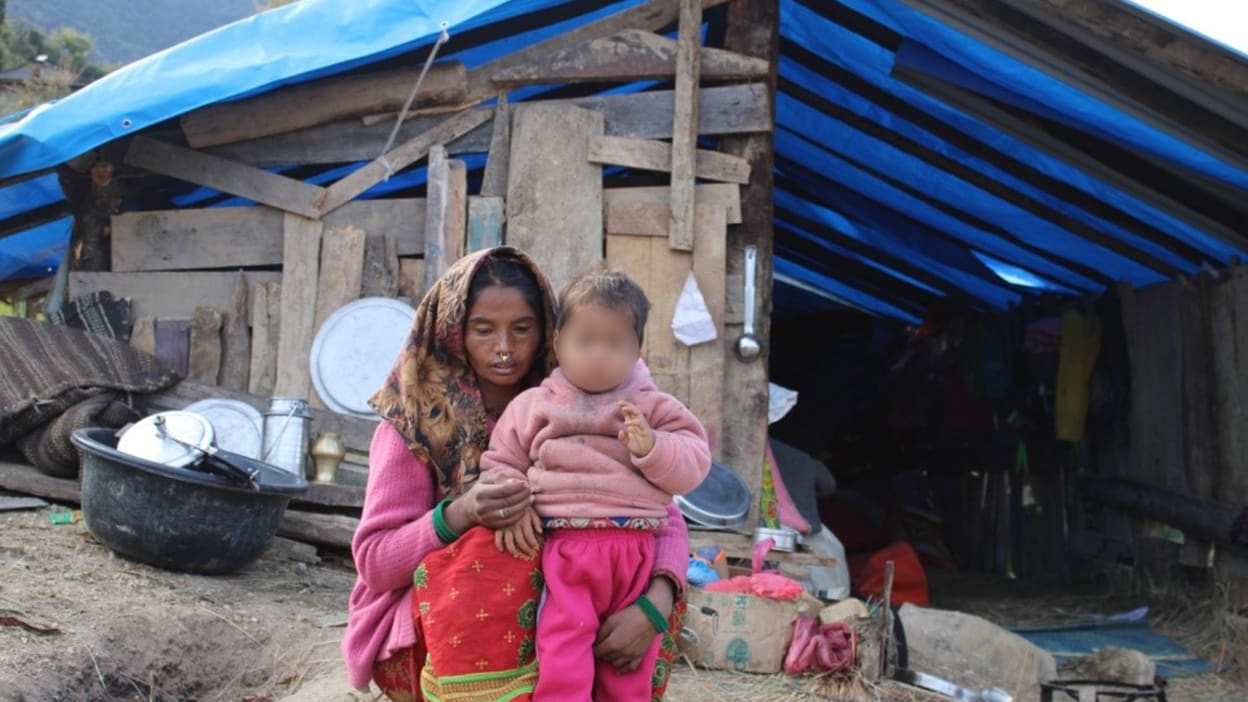 Dhanu, wearing a colourful headscarf, crouches with her young son in front of the makeshift shelter they have been living in since the earthquake destroyed their home.