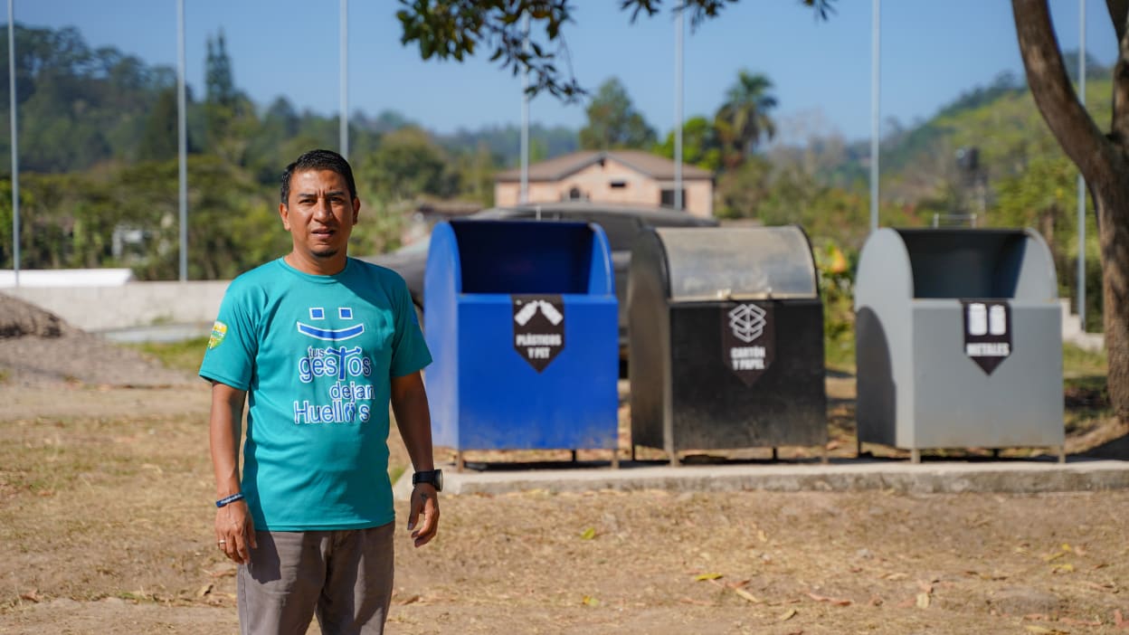 Pastor Wilfredo, dressed in a green T-shirt, stands in front of the three bins provided for waste segregation in the community.