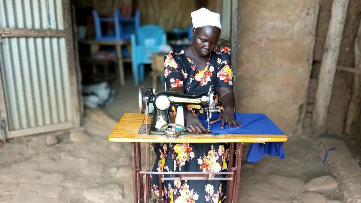 Jenifer, a young South Sudanese woman wearing a dress with bright flowers and a short headscarf, sits at a table with a sewing machine, sewing a bright blue garment.