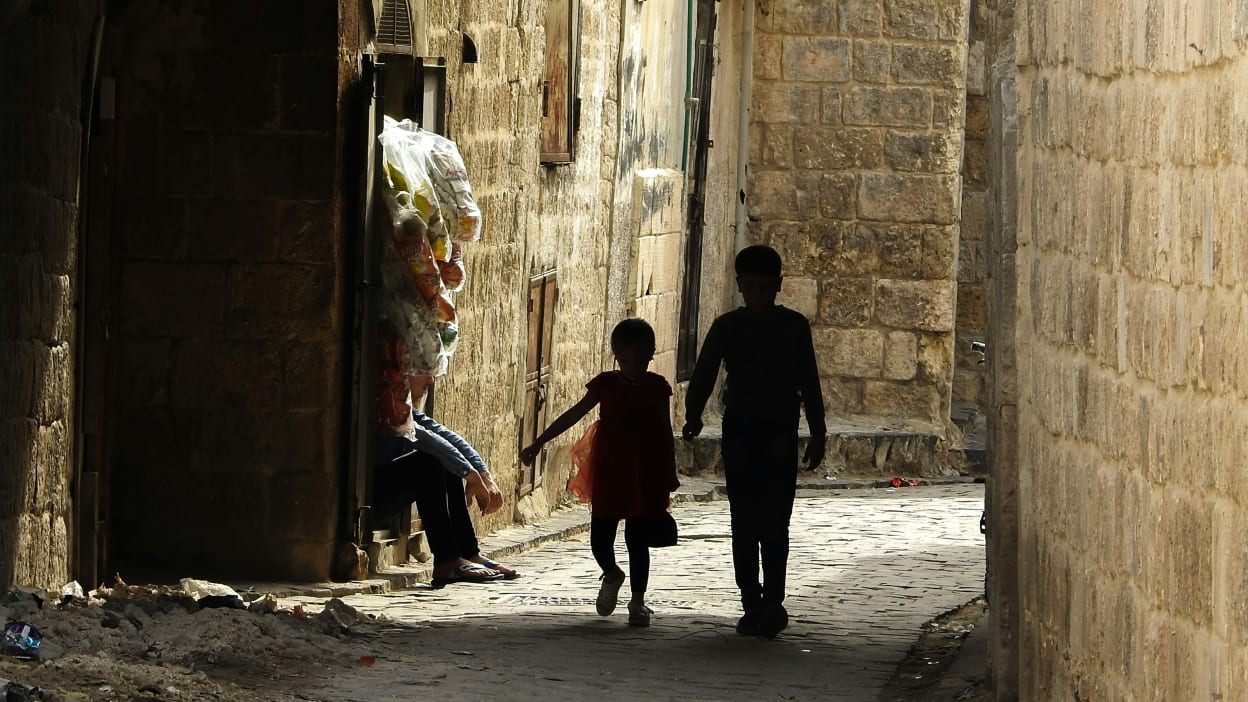 Two young children are silhouetted as they walk down a narrow alley in Aleppo, Syria. A man sits in a doorway.