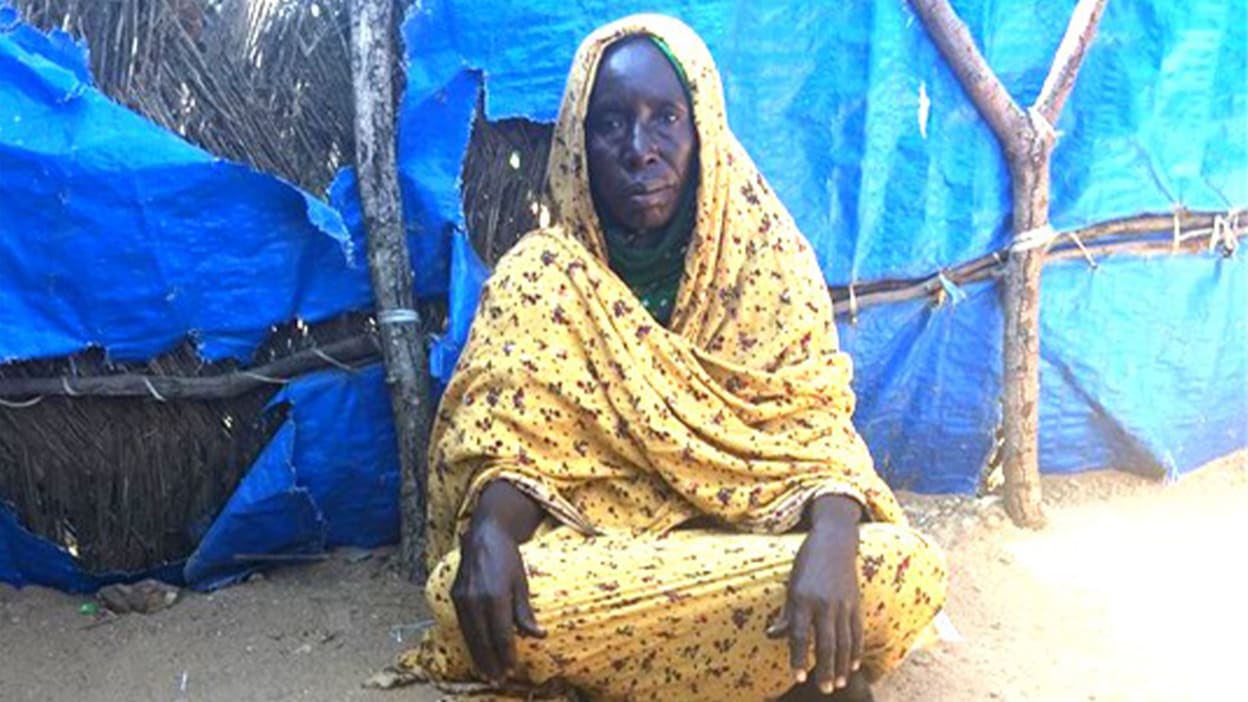 A woman is sat cross-legged on the ground in front of a wall built from wood and plastic sheets.