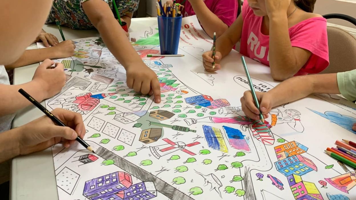 Children gathered around a table use coloured pencils to draw a town together on a large piece of paper.