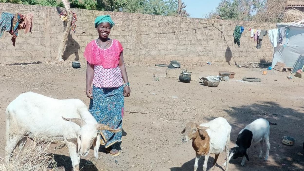 Faridatou, wearing a brightly coloured scarf on her head, smiles as she stands in her yard alongside her three short-haired sheep.