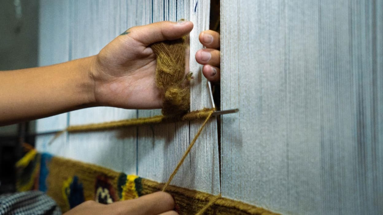 A woman's hands are shown holding a spindle with thread and weaving a carpet on a loom.