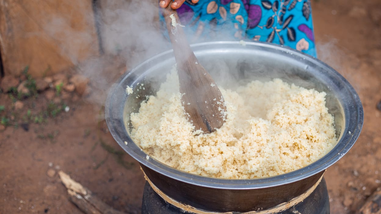 A woman in a brightly coloured dress mixes a cooking pot full of cassava couscous, called attiéké in Cote d'Ivoire
