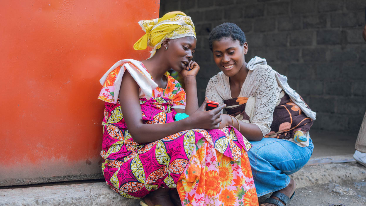 Two women sat on a step look at a phone one of the women is holding.