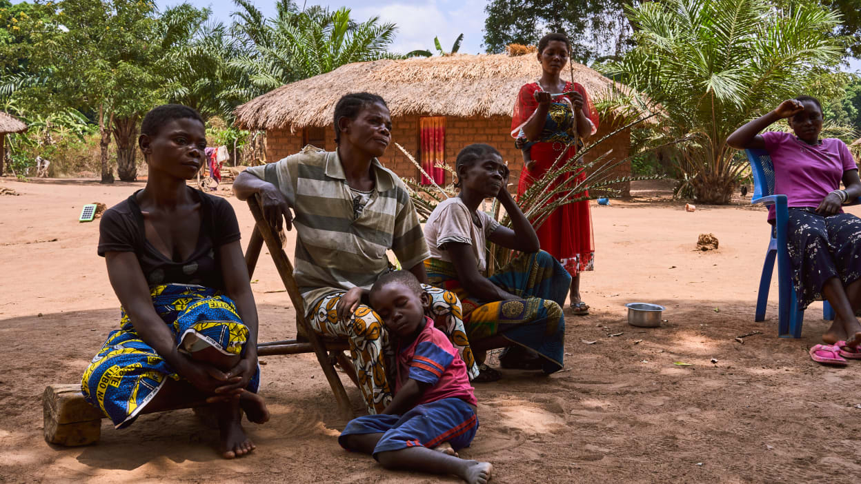 A group of woman sit outside under the shade of a tree taking part in peace dialogues. Kalonda sits with a young child leaning peacefully against her legs.