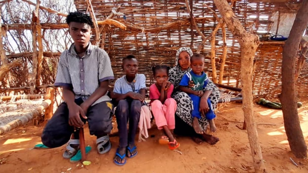 A family of five poses in front of a hut made of branches and woven wood.