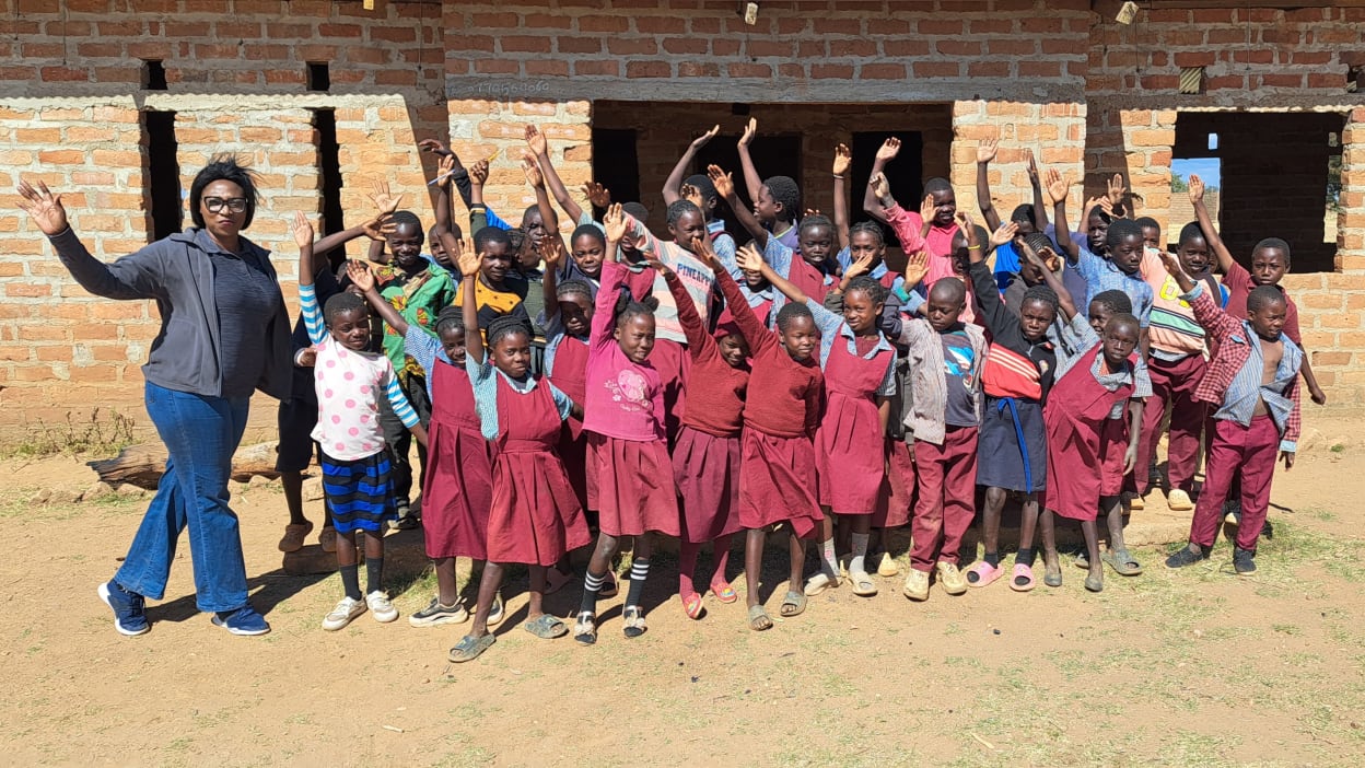 Group of African children and a woman pose with their hands up outside a brick building.