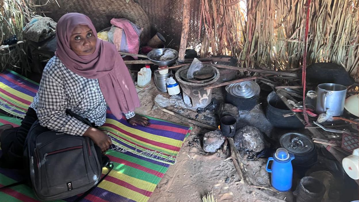 Woman in headscarf sits in a simple kitchen with cooking utensils and a straw roof.