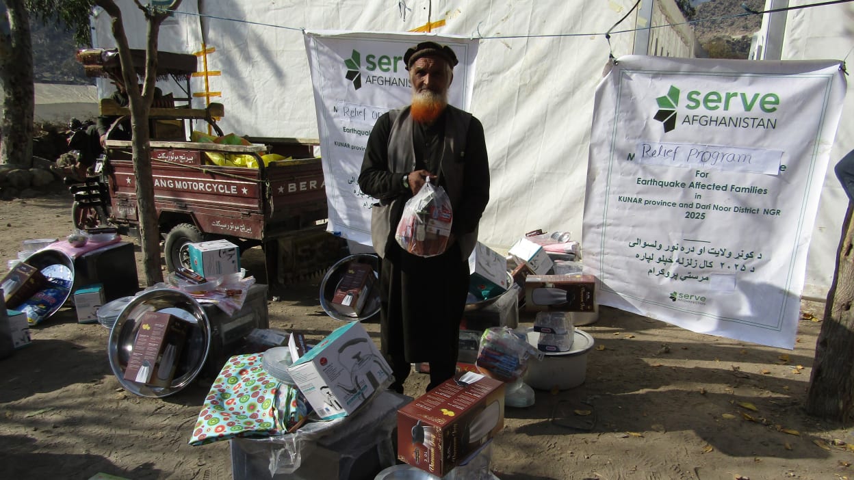 Man stands holding a bag in front of a sign that says 'Serve Afghanistan' in earthquake-affected Kunar province, Afghanistan.