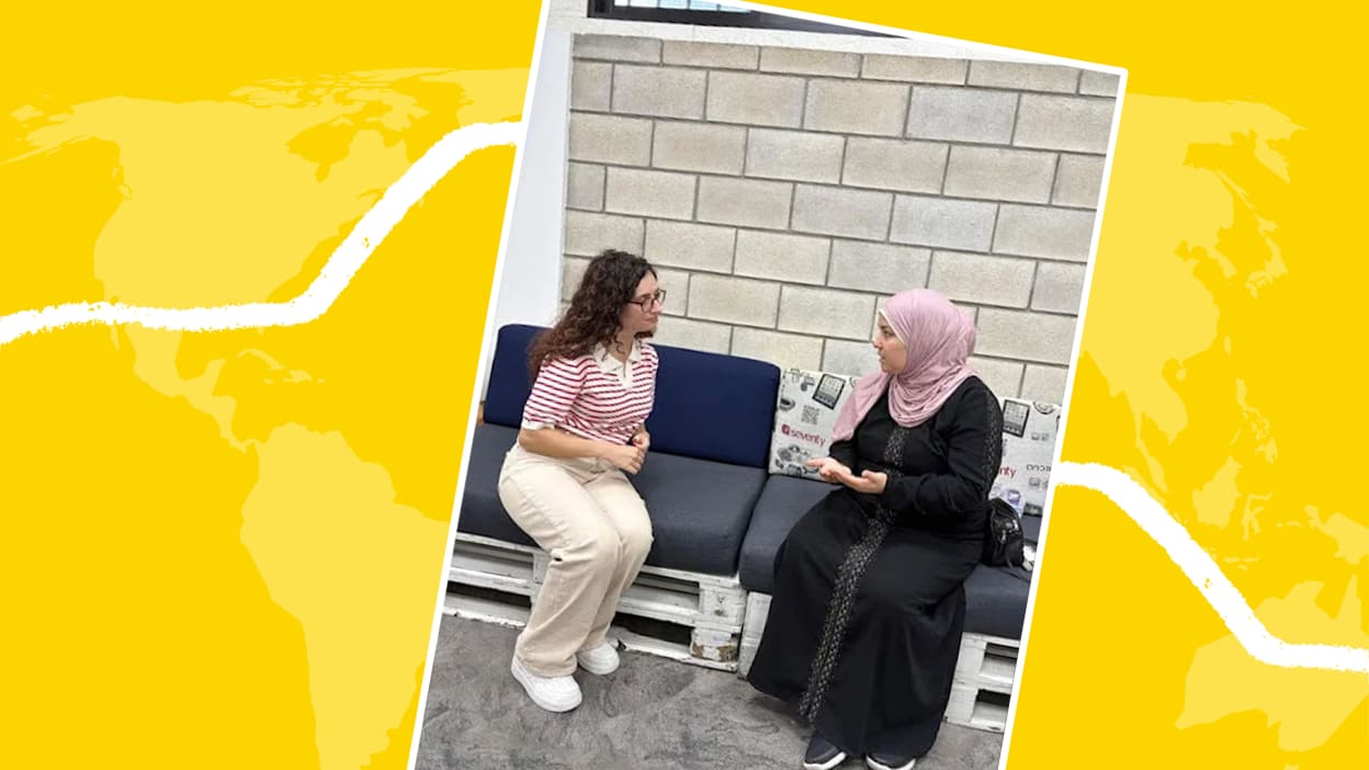 In a polaroid style graphic, a photo of two women in conversation sit on a couch against a wall with a yellow background including a world map outline.