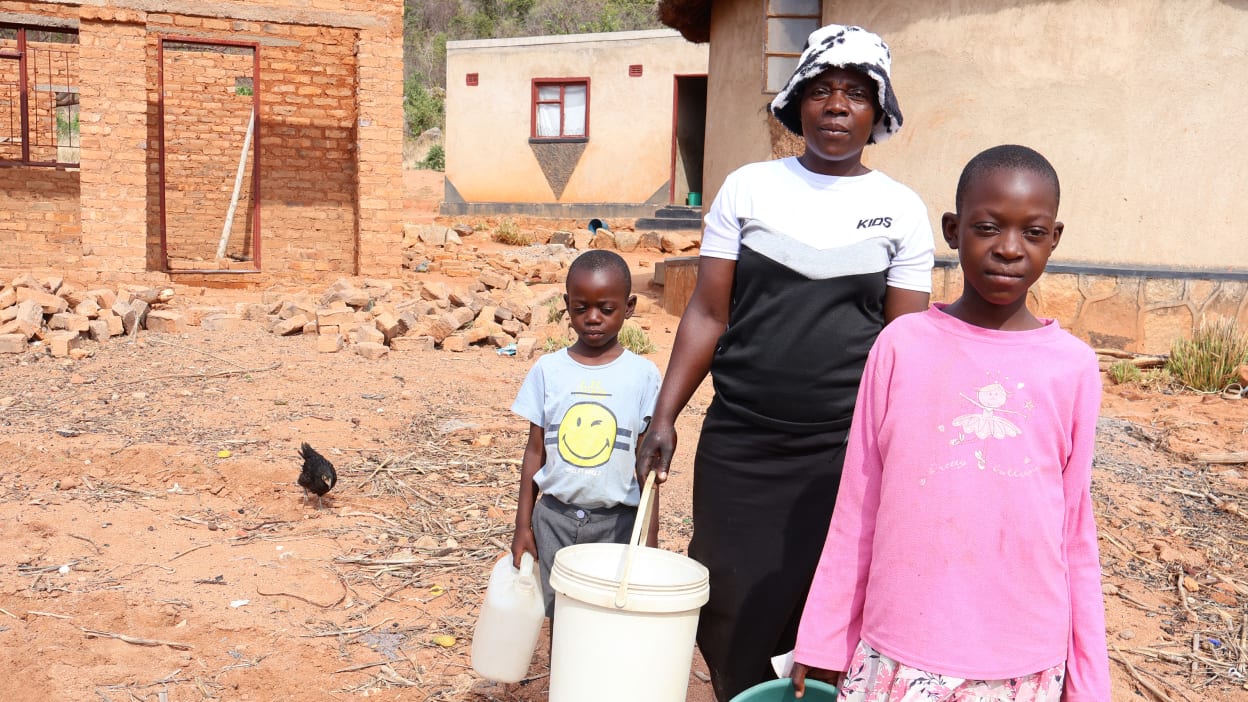 Woman and two children stand in a dusty yard with buckets, near brick buildings.