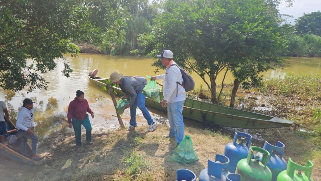 Several people stand beside a muddy riverbank and canoe, unloading green bags from the boat next to floodwater and trees.